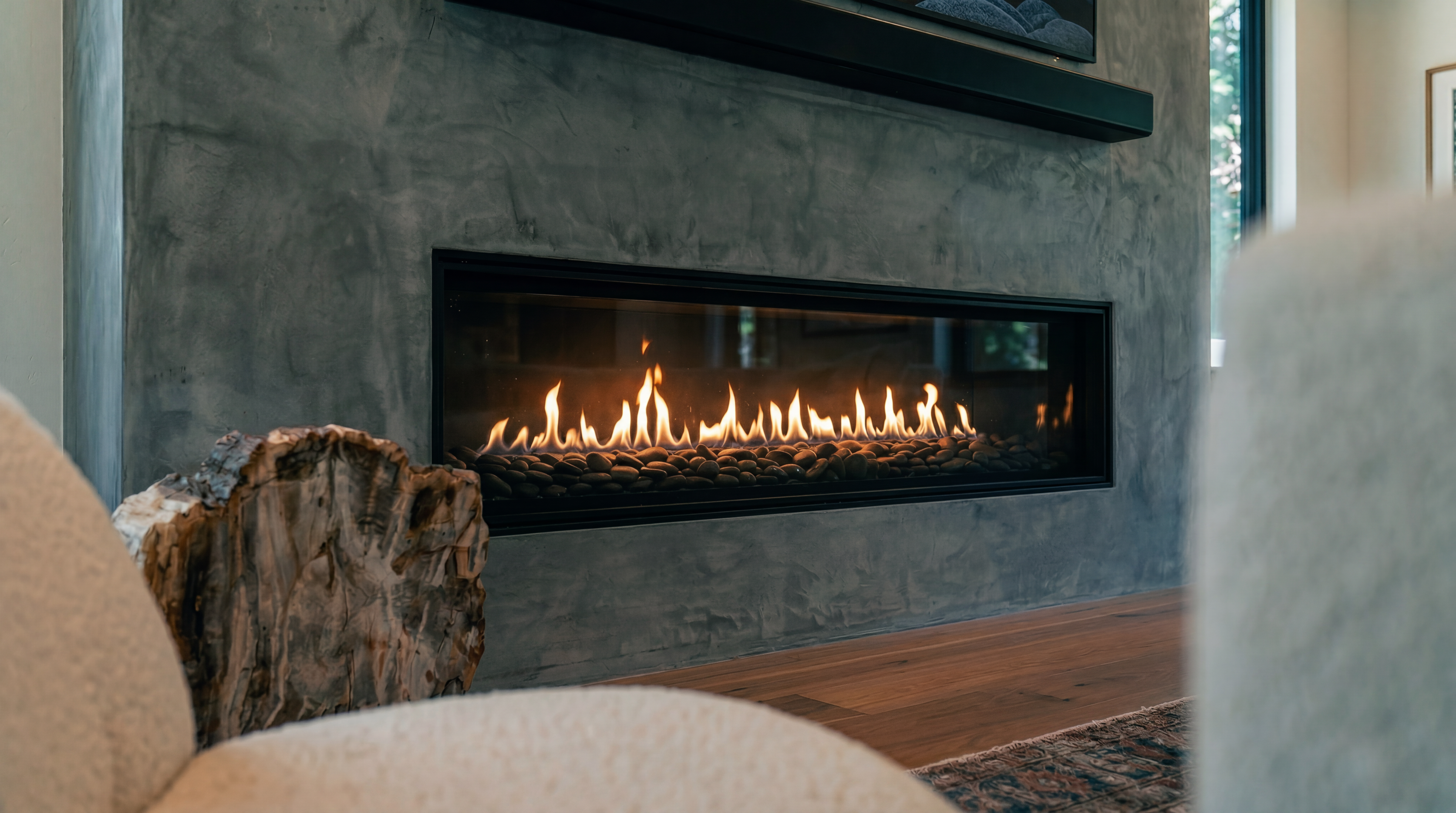 This living space features a sleek, linear gas fireplace embedded in a smooth, grey concrete-style wall, serving as a prominent focal point. The room is accented by a textured, organic stump table and plush, light-colored seating in the foreground, creating a warm and modern atmosphere. Rich hardwood flooring and a glimpse of an antique-style rug complete the elegant, cozy aesthetic of the room.