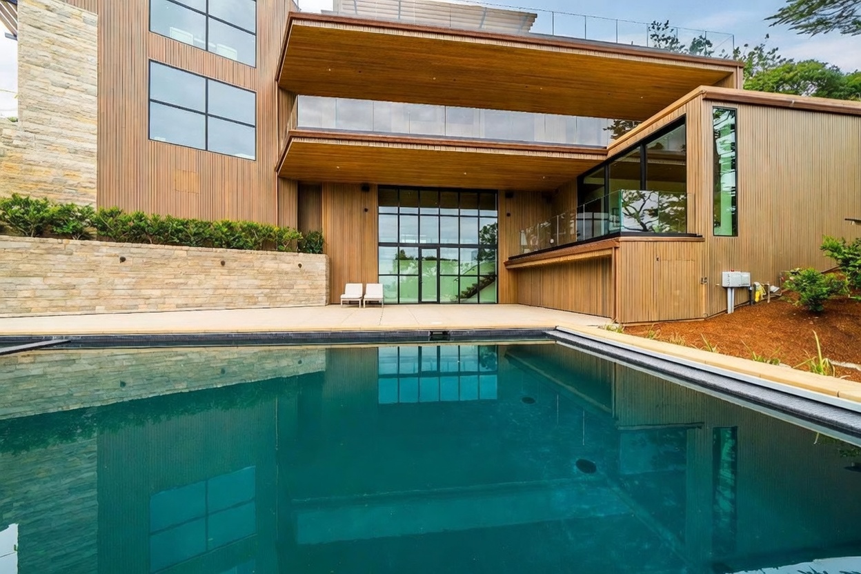 This modern architectural residence features a striking exterior with vertical wood-toned siding and extensive glass elements. A large, dark-bottomed swimming pool occupies the foreground, reflecting the home's clean lines and the surrounding landscape. The property includes a spacious patio area with two lounge chairs and a stone retaining wall, creating a sophisticated and serene outdoor living space.
