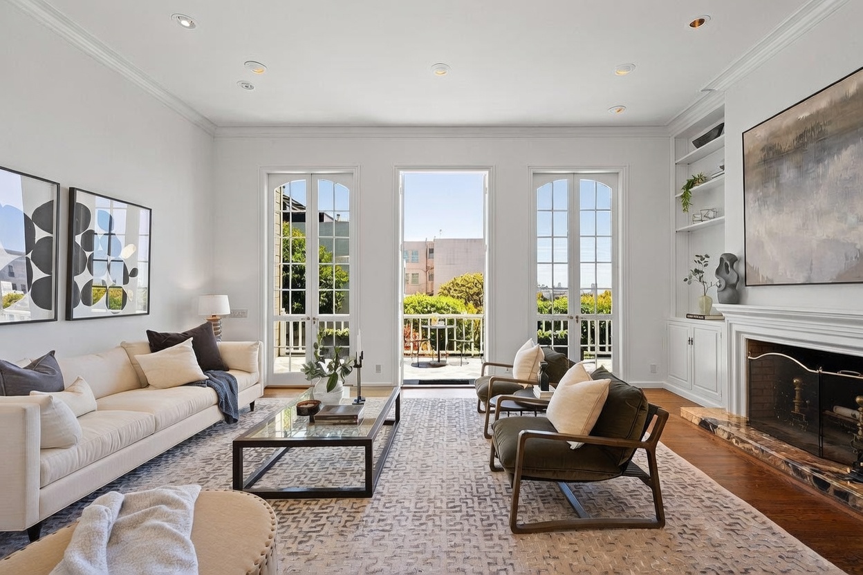 This bright and airy living room features a neutral color palette, a plush patterned area rug, and a comfortable seating arrangement centered around a glass-topped coffee table. A traditional fireplace with a white mantel serves as a focal point on the right, while three sets of tall glass doors provide abundant natural light and access to a private balcony. The space is elegantly appointed with contemporary artwork and built-in shelving, creating a sophisticated and inviting atmosphere.
