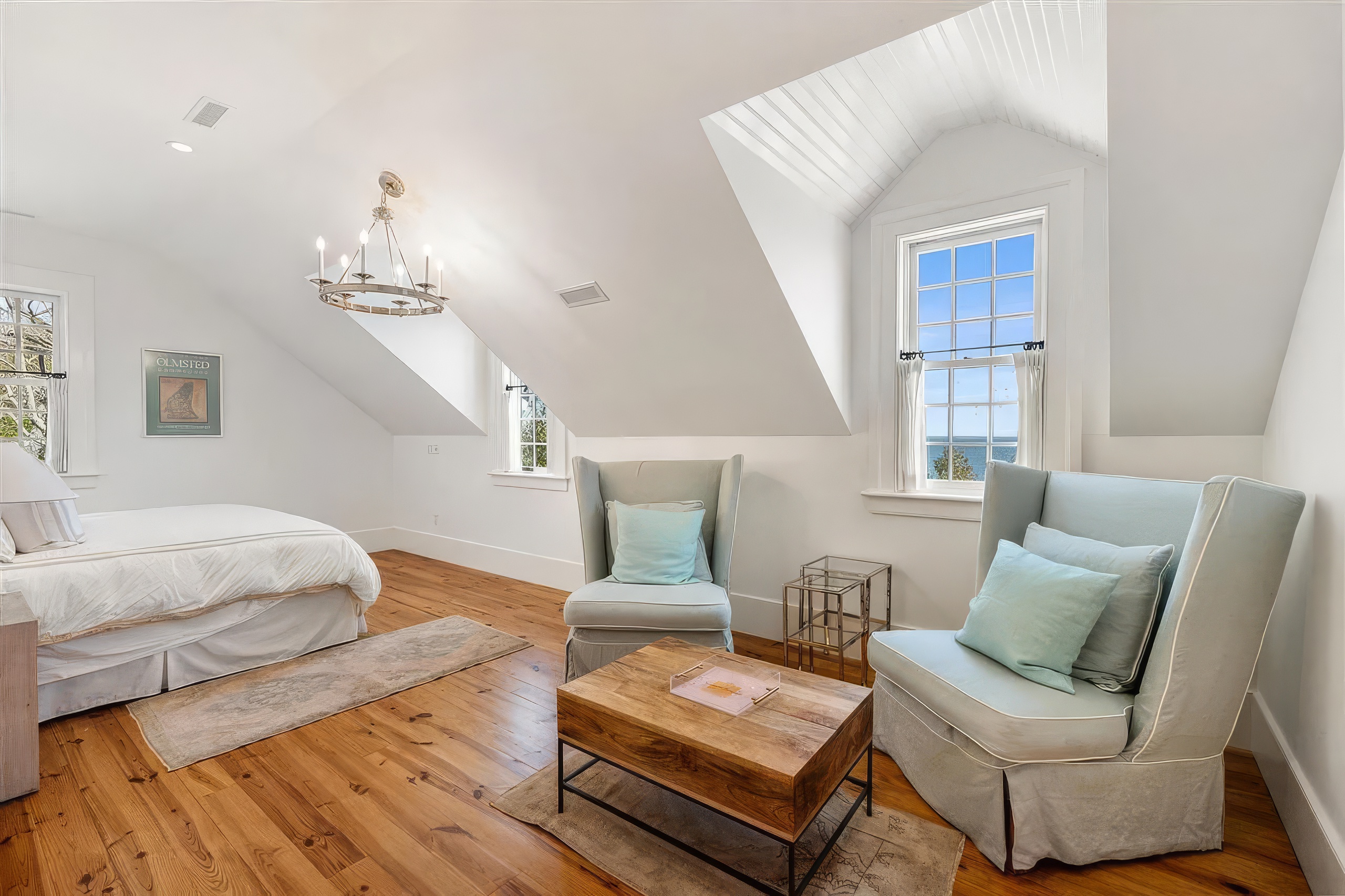 This bright and airy bedroom features a vaulted ceiling with dormer windows that offer a scenic view of the water. The room is furnished with a bed, two light-colored upholstered armchairs, and a wooden coffee table, all set upon warm-toned wood flooring. A classic chandelier hangs from the center of the ceiling, contributing to the serene and coastal-inspired aesthetic of the space.
