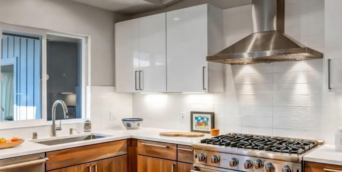 This sleek modern kitchen features a beautiful contrast between warm, wood-toned lower cabinetry and glossy, high-white upper cabinets. The workspace is equipped with a high-end stainless steel range, a professional-style range hood, and a clean white tile backsplash. A bright window above the undermount sink floods the countertop area with natural light, contributing to a polished and functional design.