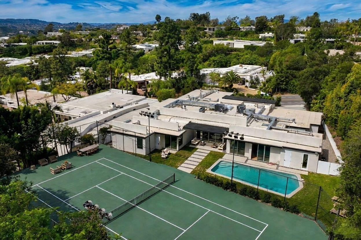 This high-angle aerial view captures a sprawling, modern single-story residence nestled in a lush, hilly landscape. The property features a private tennis court in the foreground, a rectangular swimming pool adjacent to the house, and a flat roofline punctuated by various mechanical units. The surrounding area is densely wooded with mature trees, emphasizing a secluded and expansive residential setting.