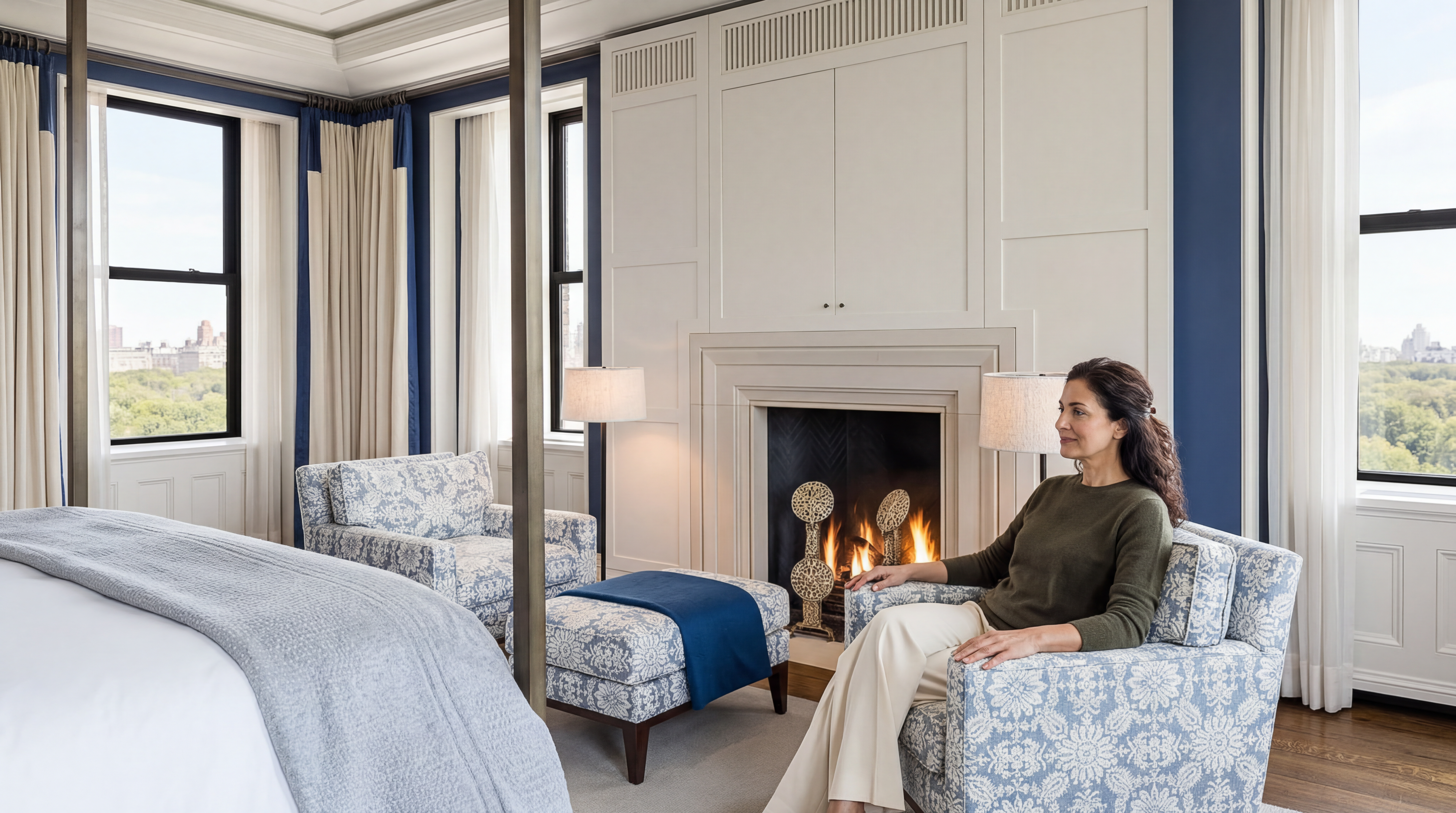 This elegant primary bedroom features a sophisticated design with a fireplace as a focal point and floor-to-ceiling accent molding. The space includes two upholstered patterned armchairs, a coordinating ottoman, and extensive views overlooking a park-filled urban skyline. The room is designed for comfort and luxury, highlighted by a four-poster bed and a warm, inviting color palette.