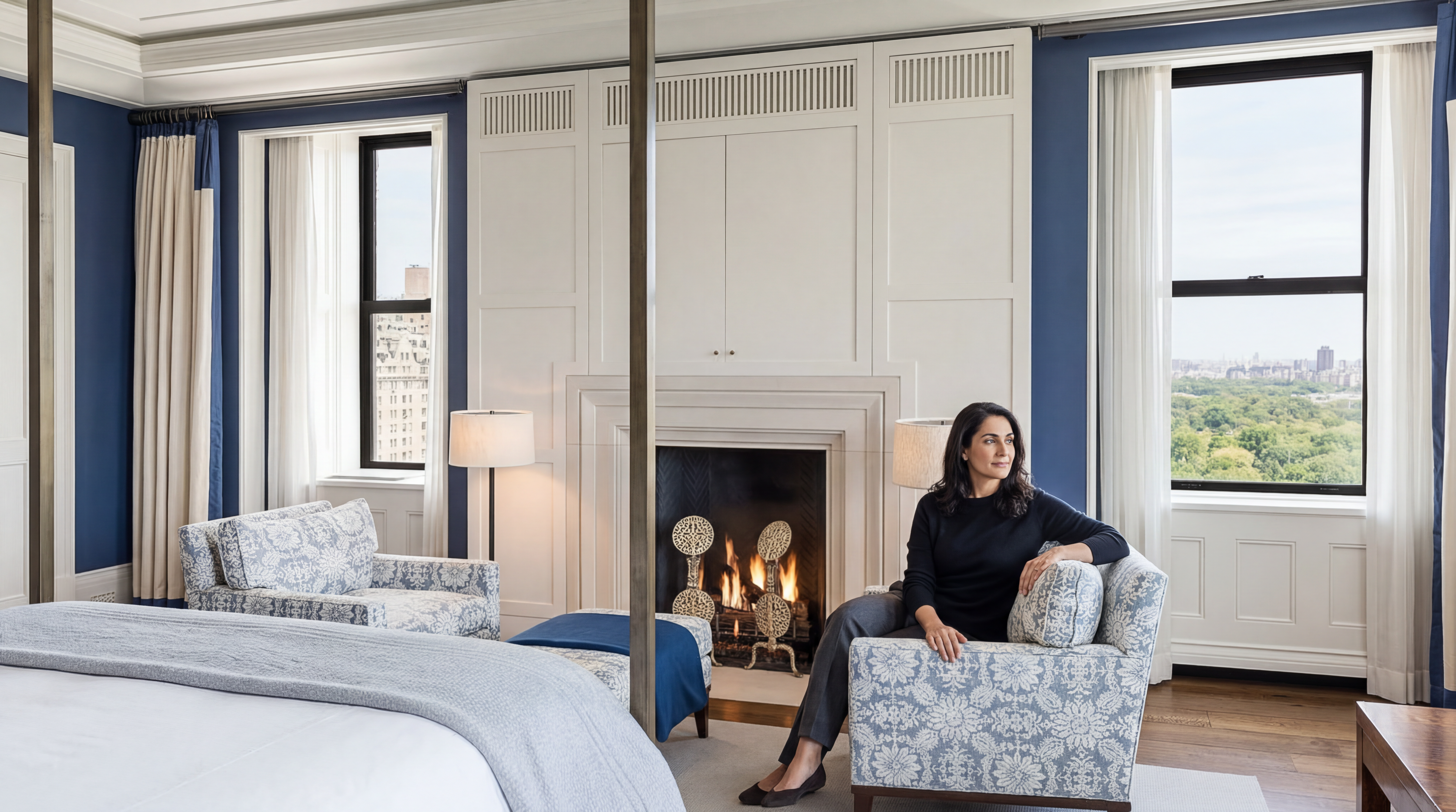 This elegant primary bedroom features a sophisticated design with a fireplace, custom millwork, and two patterned armchairs. Large windows offer breathtaking views of Central Park, bathing the space in natural light while highlighting the luxurious aesthetic and high-end finishes throughout the room.