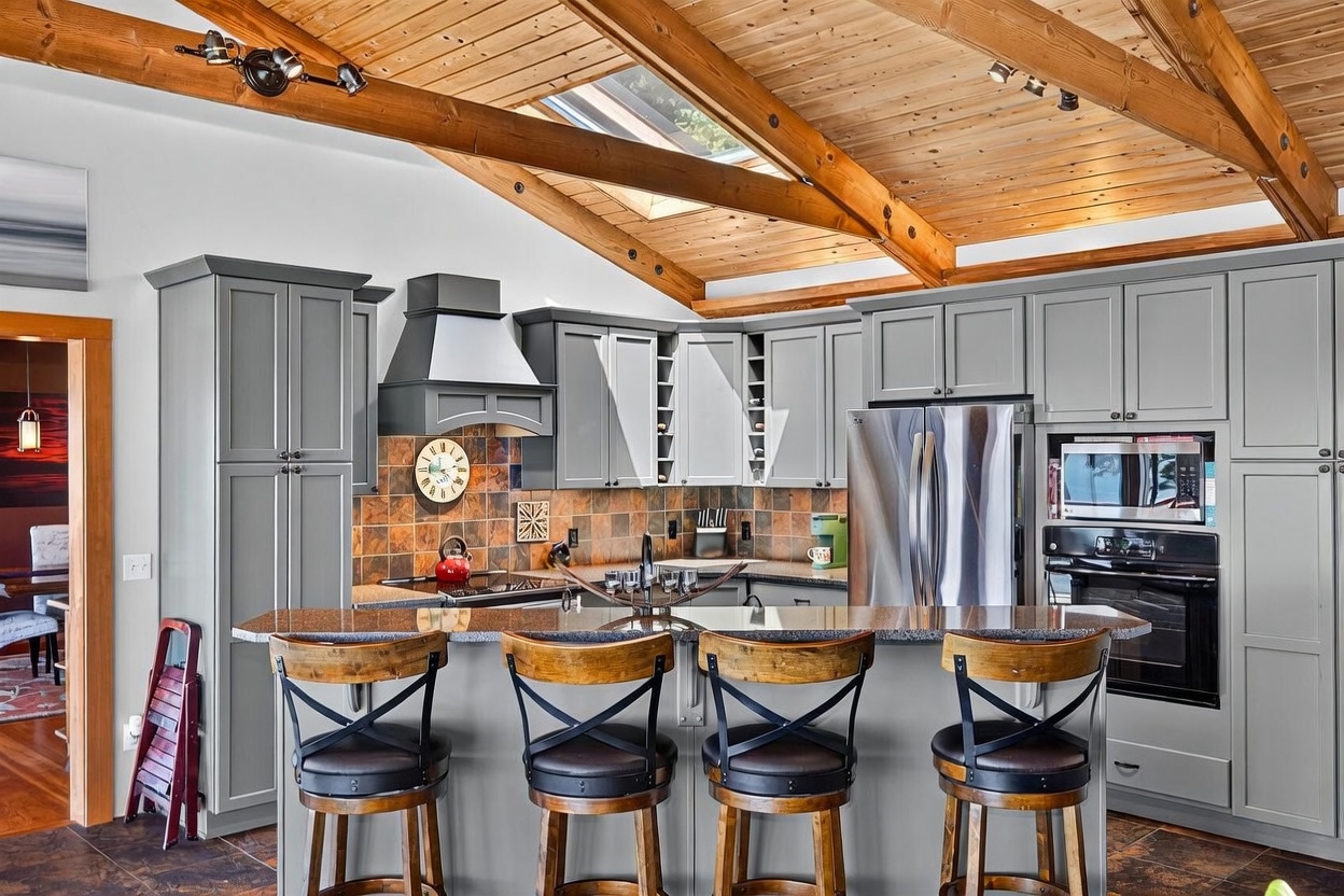 This kitchen features a vaulted wood-paneled ceiling with exposed beams and a skylight, creating an airy and rustic atmosphere. The space is equipped with grey cabinetry, a stone-tiled backsplash, and a large island with seating for four, all complemented by stainless steel appliances. The perspective is a wide-angle shot from the dining area, showcasing the open-concept layout and the warm, inviting aesthetic of the home.