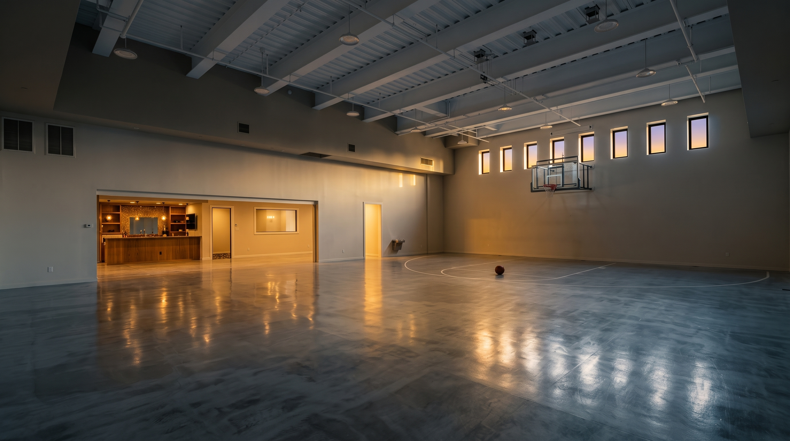 This expansive indoor sports court features a polished concrete floor and a high, exposed-beam ceiling with industrial-style lighting. A basketball hoop is mounted on the far wall, which is lined with a series of rectangular windows, while a built-in bar area with cabinetry is tucked into the side wall. The space offers a versatile, open-concept layout suitable for athletic activities or large-scale entertaining.