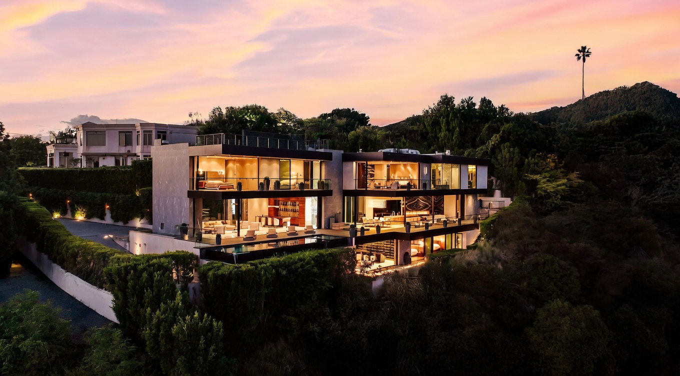 This stunning aerial view captures a contemporary multi-level luxury residence perched on a hillside at sunset. The home features expansive glass walls, multiple terraces, and a sleek infinity-edge pool that seamlessly blends with the surrounding lush landscape. The warm, golden hour lighting highlights the architectural sophistication and the dramatic, elevated setting of the property.
