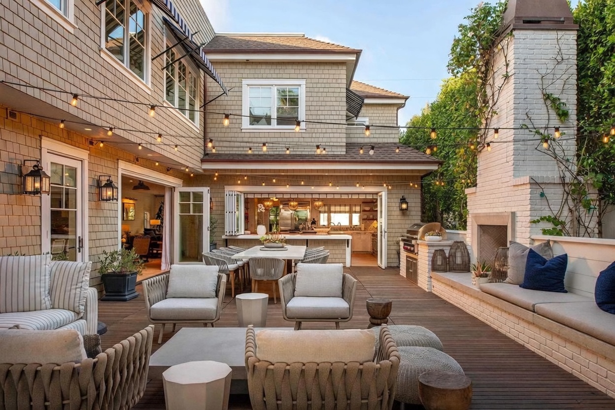 This expansive outdoor deck features a sophisticated lounge area with comfortable seating, a central dining table, and a built-in bench alongside a tall white brick chimney breast. The space seamlessly connects to the indoor kitchen through large folding doors, creating an ideal environment for alfresco entertaining. String lights are draped across the deck, enhancing the inviting atmosphere of this well-appointed backyard retreat.