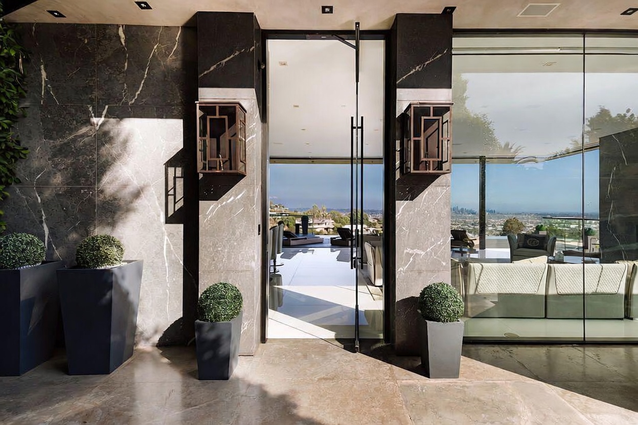 This sophisticated entryway features a grand, floor-to-ceiling glass pivot door framed by dark, polished marble walls and elegant bronze-toned wall sconces. The foreground is adorned with manicured topiary plants in sleek, modern planters, while the open door reveals a bright, expansive interior with a stunning panoramic view of the city skyline. The composition creates a seamless transition between the luxurious exterior architecture and the open-concept living space beyond.