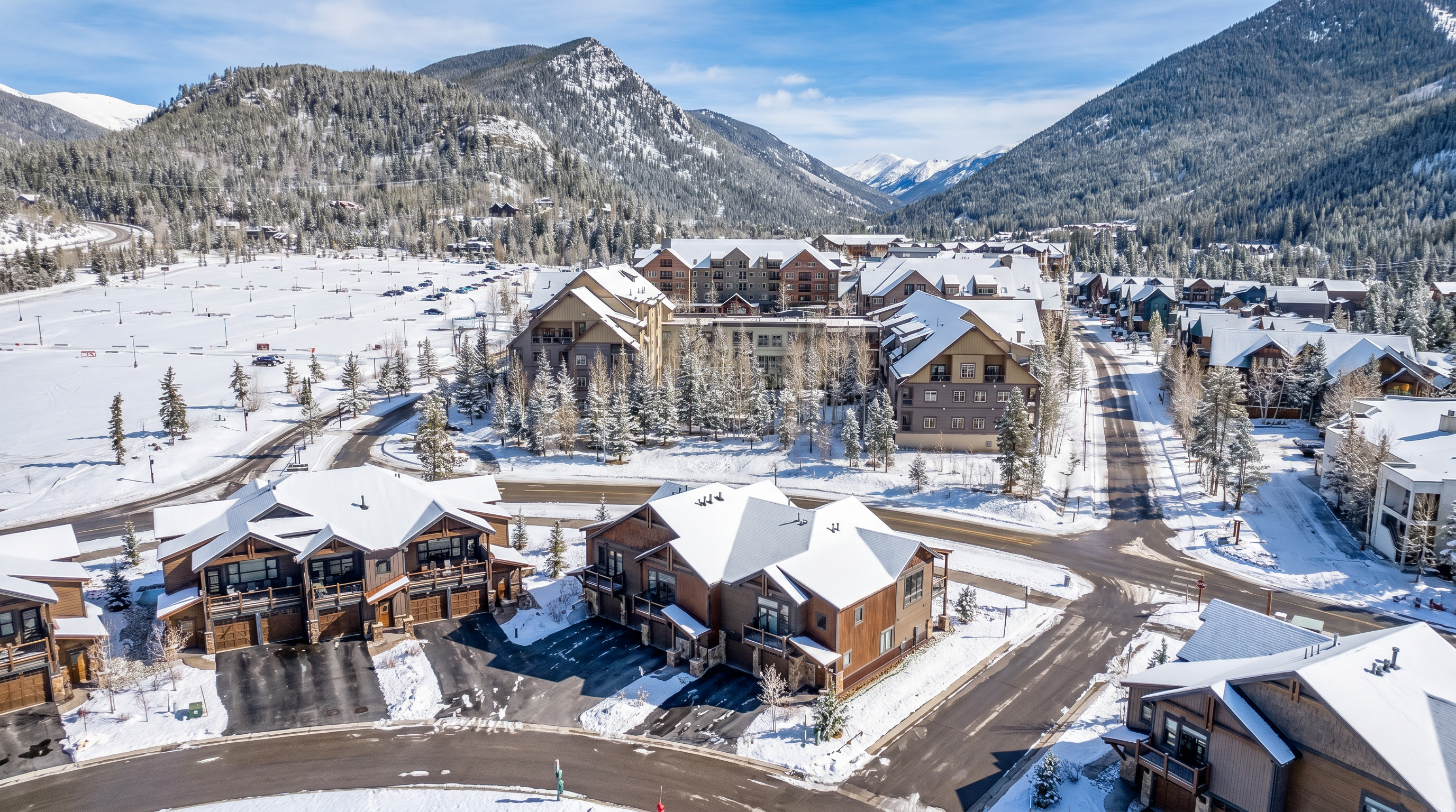 This high-angle aerial shot captures a scenic mountain resort community featuring multi-story townhomes with rustic, lodge-style architecture. The scene showcases a blend of paved roads, landscaped grounds, and a large parking area, all set against a backdrop of majestic, snow-dusted mountain peaks under a clear blue sky. The perspective provides a comprehensive overview of the neighborhood's layout and its integration into the surrounding natural landscape.