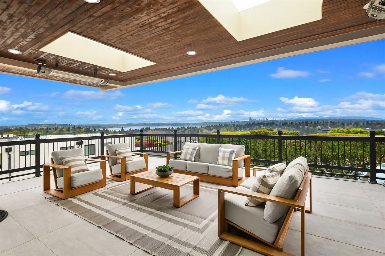 This expansive covered balcony offers a sophisticated outdoor living space with a wood-paneled ceiling, recessed lighting, and a skylight. The area is furnished with a modern light wood seating set, including a sofa and armchairs arranged around a central coffee table on a patterned area rug. A black metal railing frames the space, providing an unobstructed, panoramic view of the surrounding landscape, distant water, and city skyline.