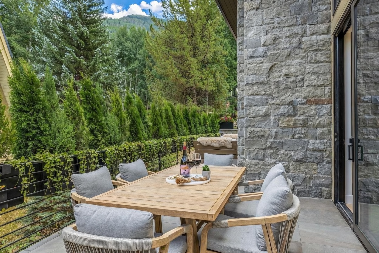 This outdoor patio space features a light wood dining set with cushioned chairs, perfect for al fresco dining against a backdrop of lush evergreen trees and mountain views. The area is defined by a textured stone wall and a modern metal railing draped with climbing greenery, creating a private and serene atmosphere. The perspective captures the inviting setup from a slightly elevated angle, emphasizing the seamless transition between the indoor living space and the natural surroundings.