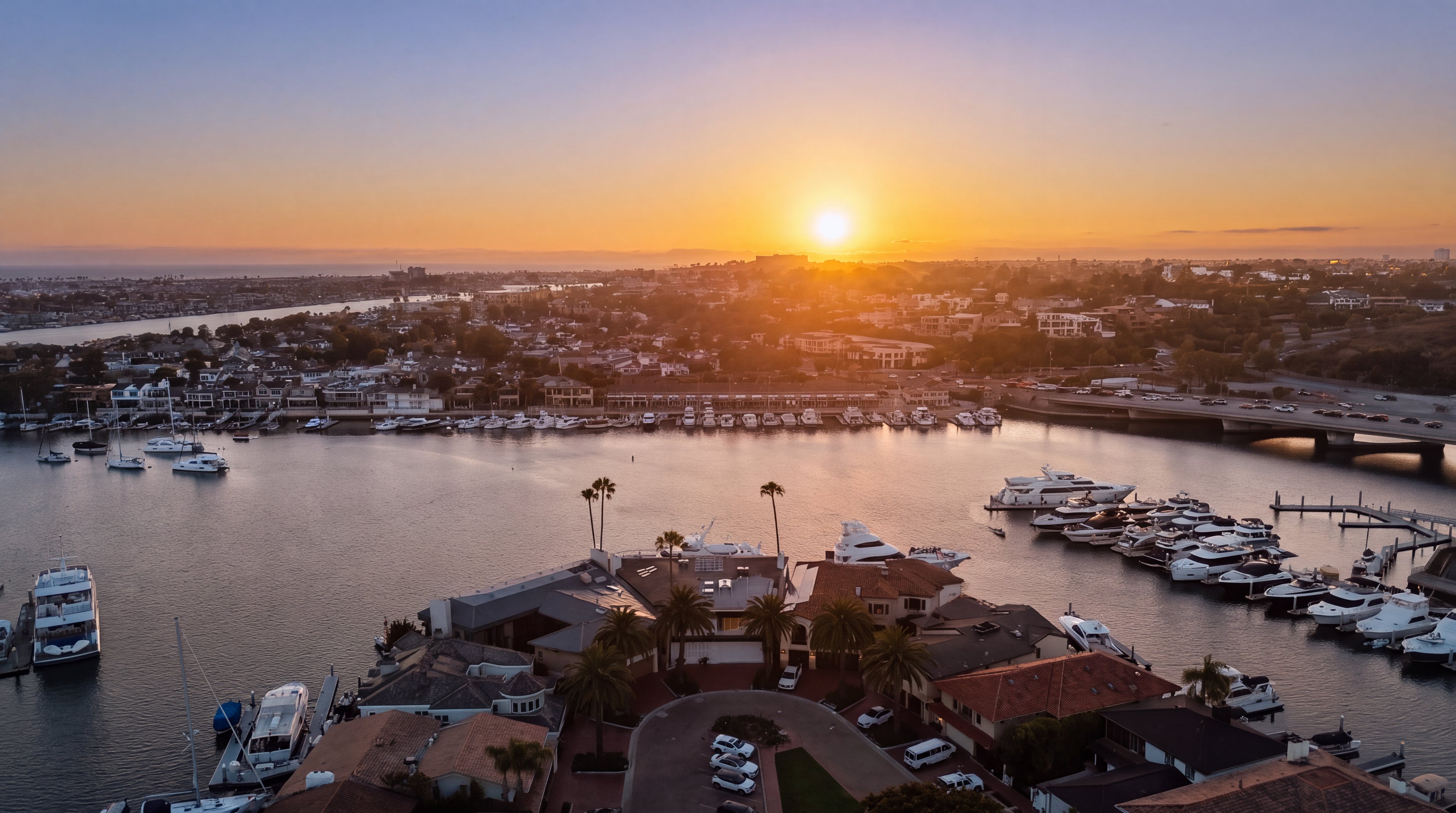 This stunning aerial view captures a waterfront residential property at sunset, showcasing its prime location along a bustling harbor filled with numerous yachts and boats. The perspective highlights the expansive water views, the surrounding neighborhood layout, and a nearby bridge, all bathed in the warm, golden light of the setting sun. The scene emphasizes a luxurious coastal lifestyle with direct water access and panoramic vistas.