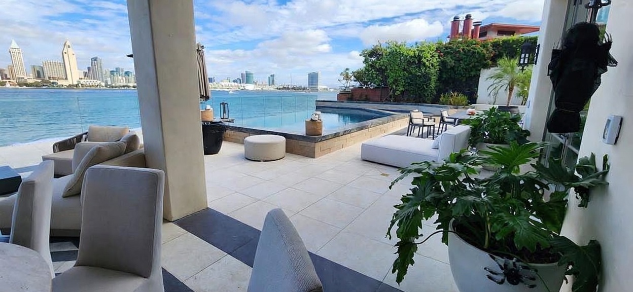 This luxurious waterfront patio offers a seamless transition between indoor and outdoor living, featuring light-toned stone flooring and modern lounge furniture. The space overlooks a private infinity-edge pool that visually merges with the expansive bay and the distant city skyline. The setting is sophisticated and serene, perfect for high-end entertaining with a clear, unobstructed view of the water.