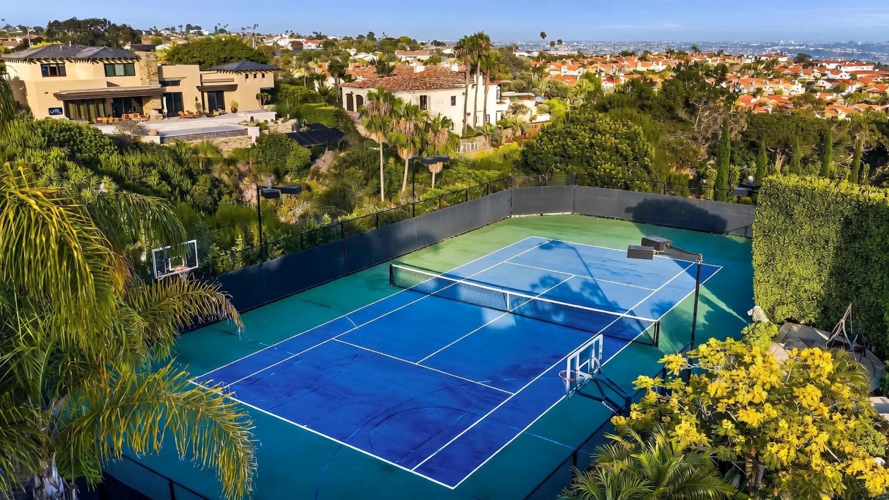 This high-angle aerial shot captures a private tennis court with a basketball hoop, set against a lush, hilly landscape. In the background, a large, multi-level residence with a contemporary design overlooks the court, surrounded by mature trees and a sprawling suburban neighborhood. The perspective emphasizes the expansive outdoor recreational space and the property's elevated position within the community.