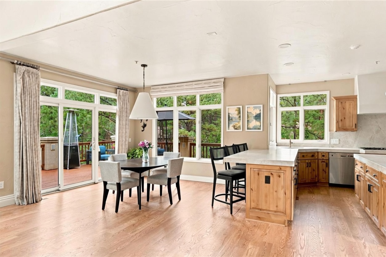 This open-concept kitchen and dining area features light wood cabinetry, a large central island with a stone countertop, and a dedicated dining space with a modern pendant light. Large windows and sliding glass doors provide abundant natural light and views of the surrounding trees and outdoor deck. The space is finished with light-toned hardwood flooring, creating a bright and inviting atmosphere suitable for both cooking and entertaining.