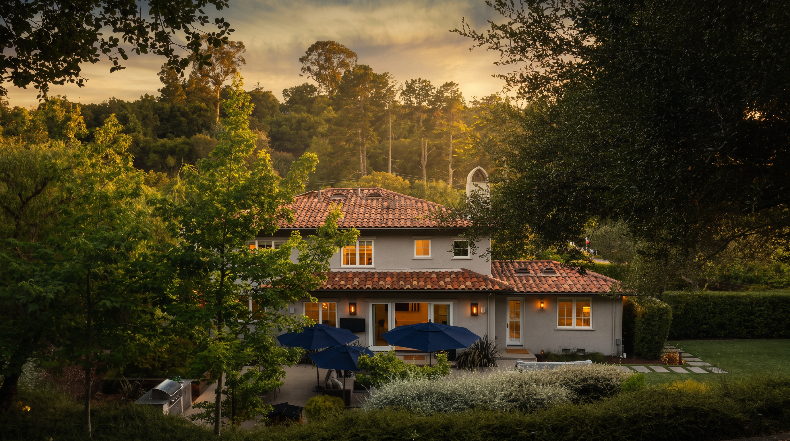 This rear view of a two-story residence showcases a Mediterranean-inspired architectural style with a classic terracotta tile roof and light-colored stucco exterior. The property features an inviting backyard patio equipped with multiple blue umbrellas for outdoor dining and lounge space, nestled amongst lush trees and well-maintained greenery. The setting offers a peaceful and private atmosphere perfect for outdoor entertaining.