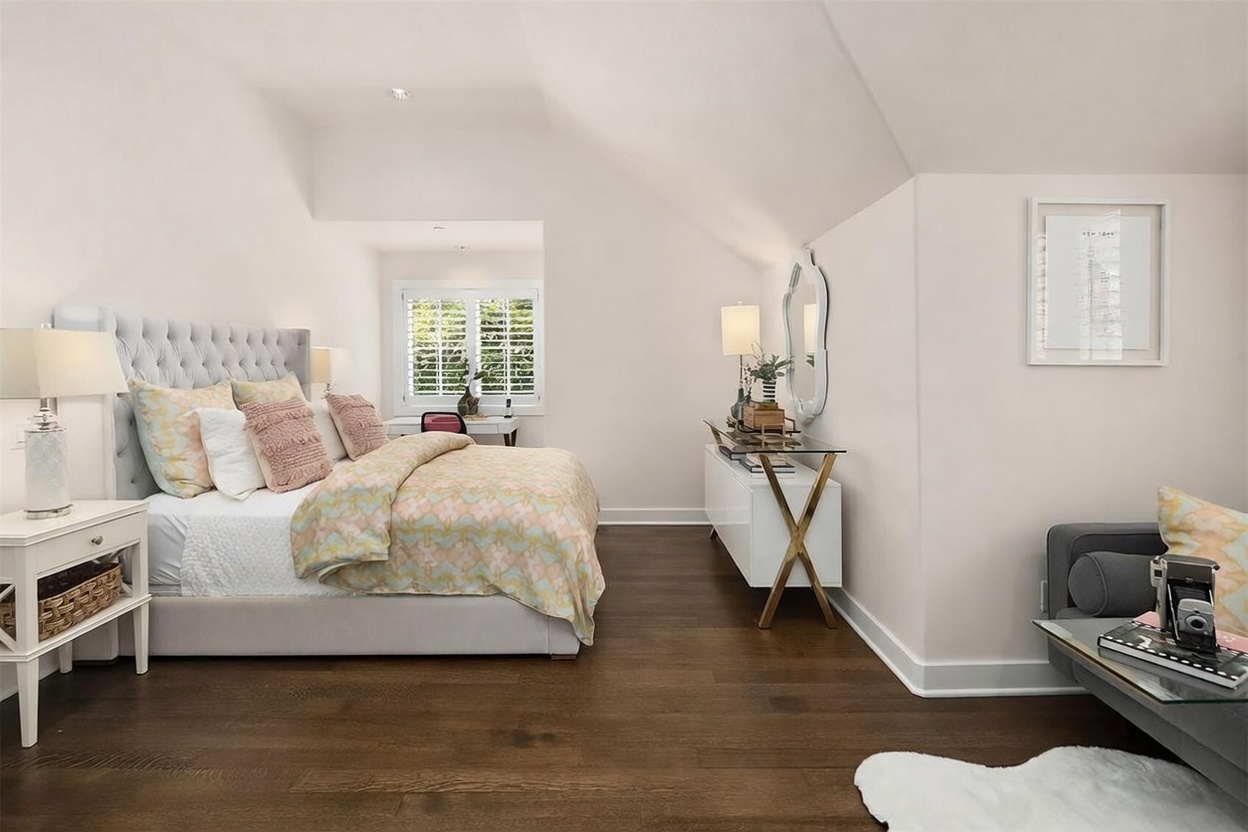 This bright and airy primary bedroom features a tufted upholstered headboard, a patterned duvet, and dark wood flooring. The room includes a sleek white console table with a decorative mirror, a small desk area by the window with plantation shutters, and a framed map of New York on the wall. The perspective is from the doorway, looking into the bedroom to showcase the layout and elegant decor.