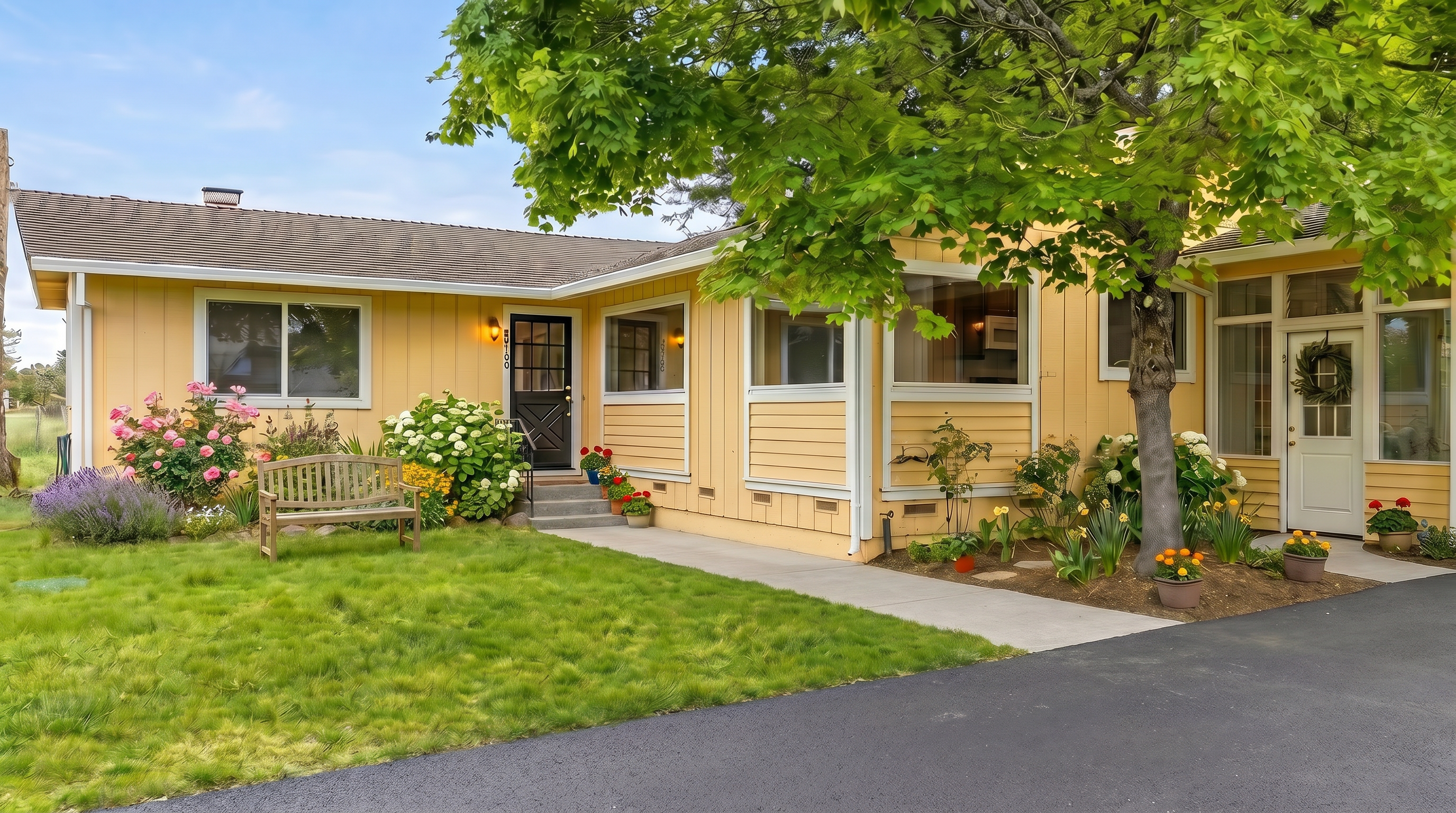 This single-story home features a light yellow exterior with a mix of horizontal siding and large windows that glow warmly in the twilight. The front entrance is accessed by a concrete walkway leading to a dark-colored door, while a mature tree provides shade over the right side of the house. A gravel driveway sits in the foreground, and the overall impression is a welcoming, well-lit residential property at dusk.