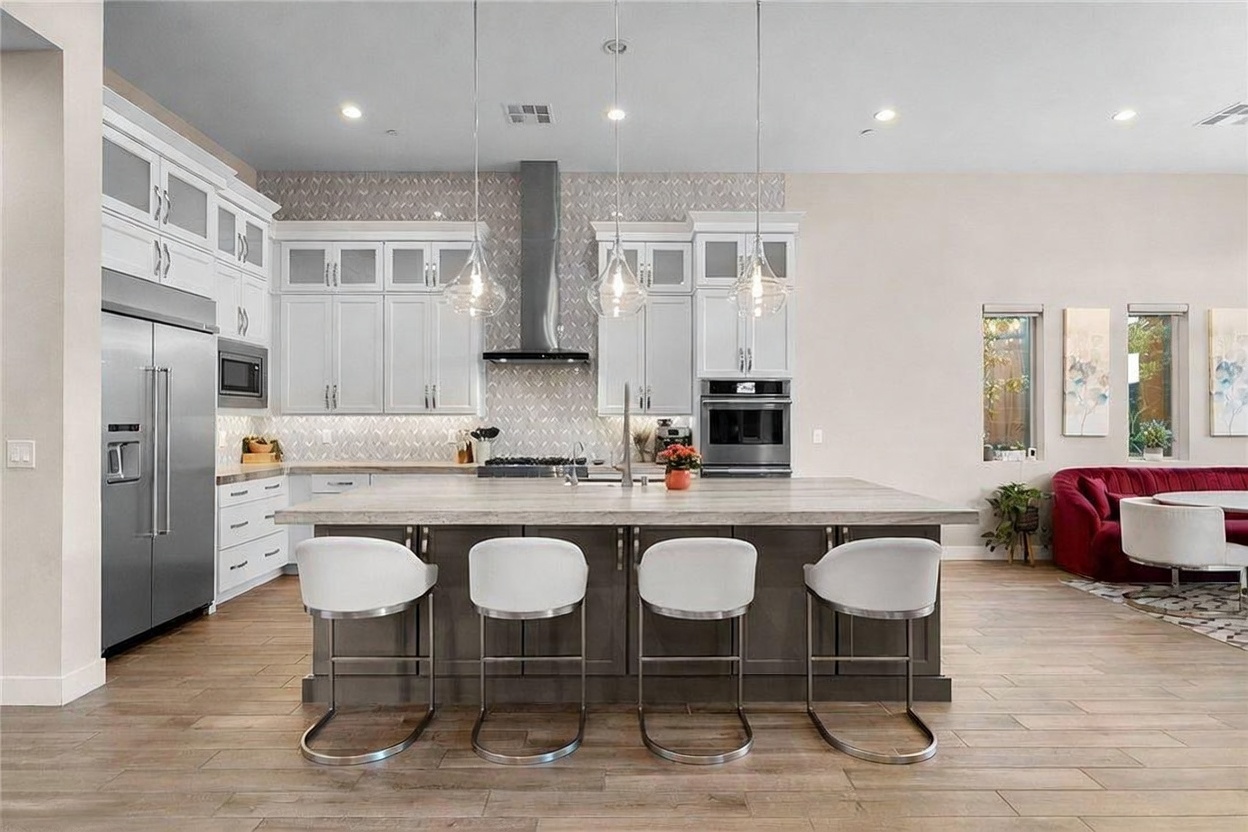 This modern kitchen features a large central island with a stone countertop and four white bar stools, serving as the focal point of the open-concept space. The room is equipped with white cabinetry, stainless steel appliances, and a patterned tile backsplash, all illuminated by three glass pendant lights. To the right, a glimpse of a dining area with a red velvet bench adds a touch of color to the neutral, light-toned wood flooring.
