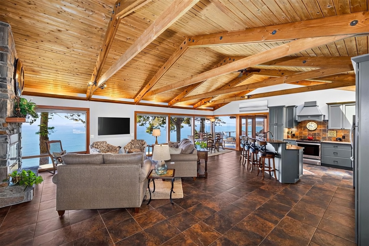 This open-concept living area features a vaulted wood-paneled ceiling with exposed beams and dark tile flooring throughout. A stone-clad chimney breast anchors the space, while large windows provide expansive views of the ocean and surrounding trees. The room flows seamlessly into a kitchen with dark cabinetry and a breakfast bar, creating a bright and airy atmosphere.