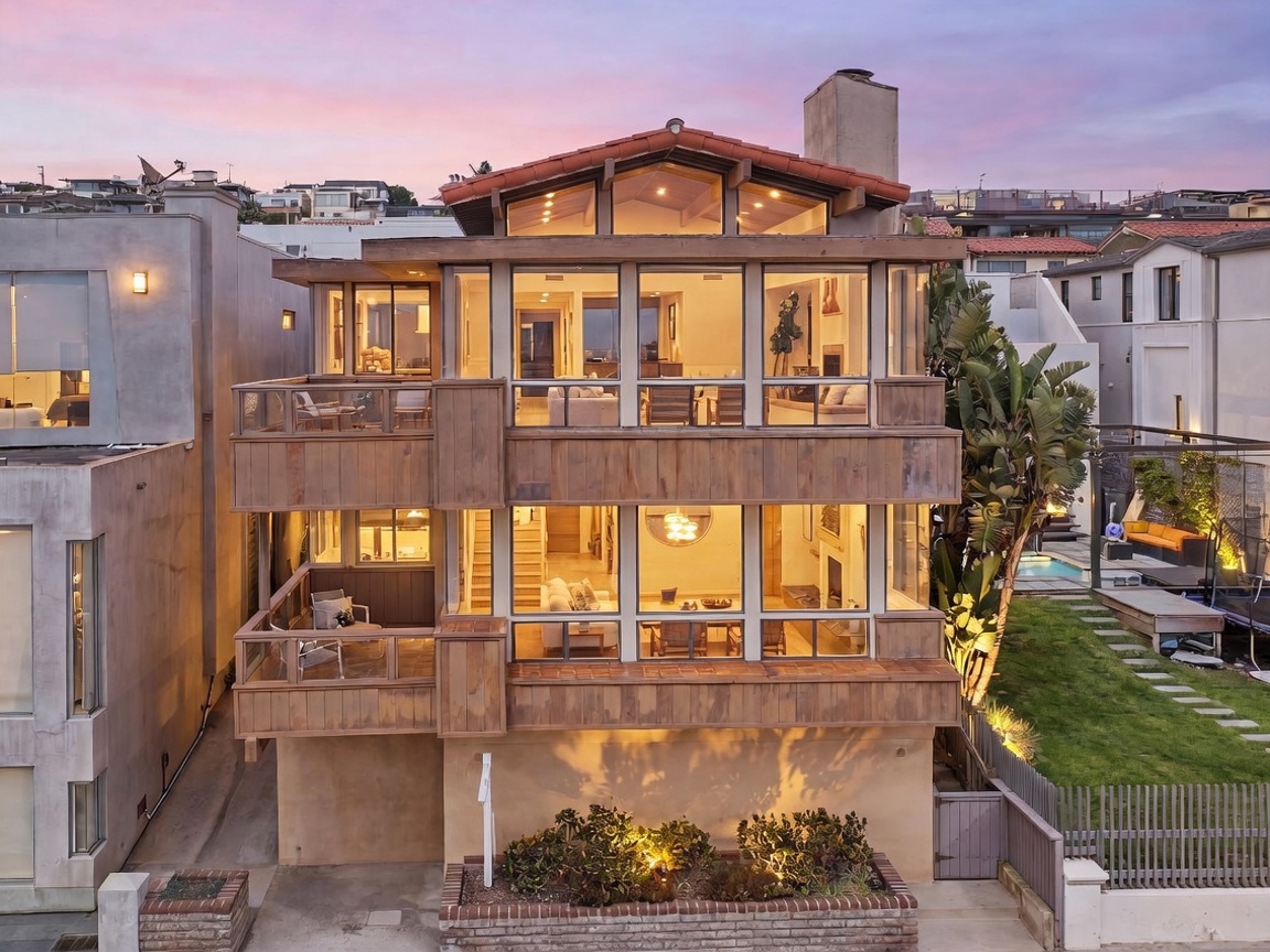 This multi-level contemporary residence features a striking facade with extensive glass glazing across the upper floors, offering an open and airy aesthetic. The exterior is finished with warm-toned wood siding and a tiled roof, complemented by a landscaped front garden and a private balcony on the second level. The perspective is a straight-on exterior shot captured at dusk, highlighting the warm interior lighting that glows through the large windows.