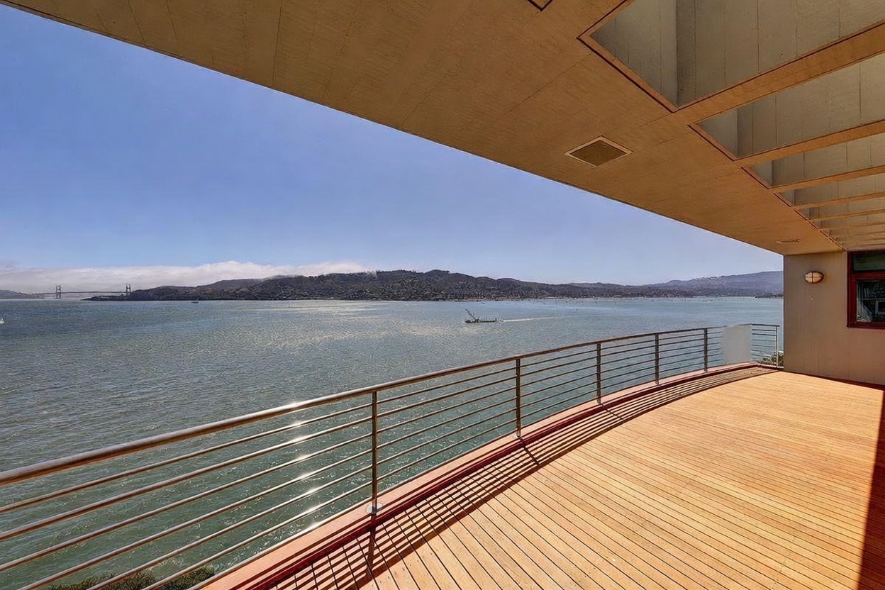 This expansive, curved wooden deck offers a panoramic, elevated view of a large bay with distant hills and a bridge visible on the horizon. The space is defined by a sleek metal railing and a deep, covered overhang that provides shade while framing the scenic waterfront vista. The perspective is from the deck looking outward, emphasizing the seamless connection between the home's outdoor living area and the natural landscape.