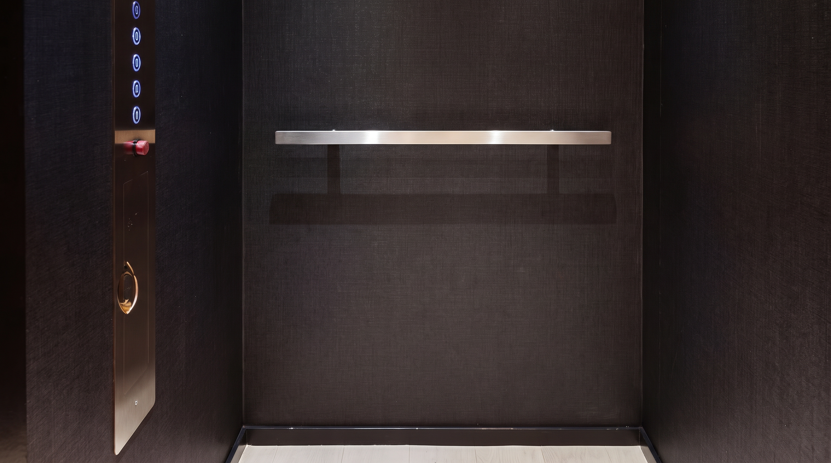This image shows the interior of a modern, minimalist elevator cabin featuring dark, textured wall panels and a light-toned wood floor. A sleek, horizontal metal handrail is mounted on the back wall, and a vertical control panel with illuminated buttons is positioned on the left side. The space is illuminated by two recessed ceiling lights, creating a sophisticated and clean aesthetic.