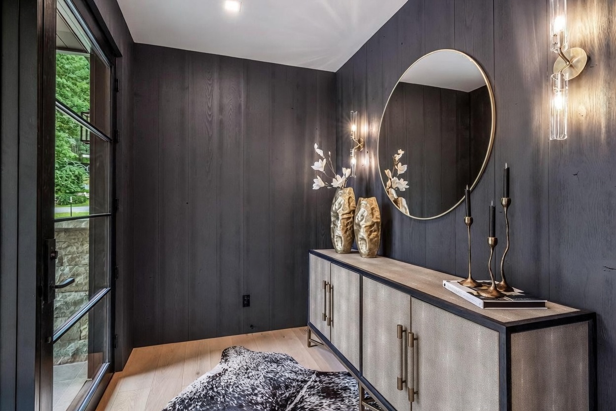 This sophisticated entryway features dramatic dark wood-paneled walls that create a moody, high-end atmosphere. A sleek console table with textured doors and metallic hardware sits beneath a large circular mirror, complemented by elegant wall sconces and decorative vases. The space is grounded by light wood flooring and a patterned animal-hide rug, while a glass-paned door provides a view of the lush greenery outside.