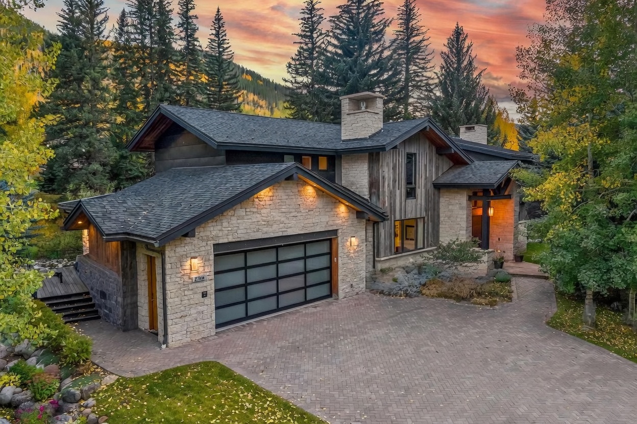 This striking mountain-style residence features a sophisticated blend of light-toned stone masonry and vertical wood siding, accented by dark trim and a multi-gabled roofline. The property includes a spacious paved driveway leading to a modern, multi-panel glass garage door, while the surrounding landscape is filled with mature evergreen and aspen trees. The perspective is an elevated, wide-angle shot that captures the home's architectural integration with its natural, wooded environment during a vibrant sunset.