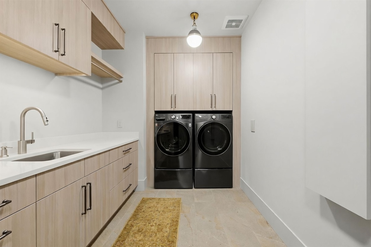 This modern laundry room features light wood cabinetry, a sleek white countertop with an integrated sink, and a pair of dark-toned front-load washer and dryer units. A textured runner rug adds warmth to the stone-tiled floor, while a single pendant light provides focused illumination. The space is clean and functional, offering ample storage with both upper cabinets and lower drawers.