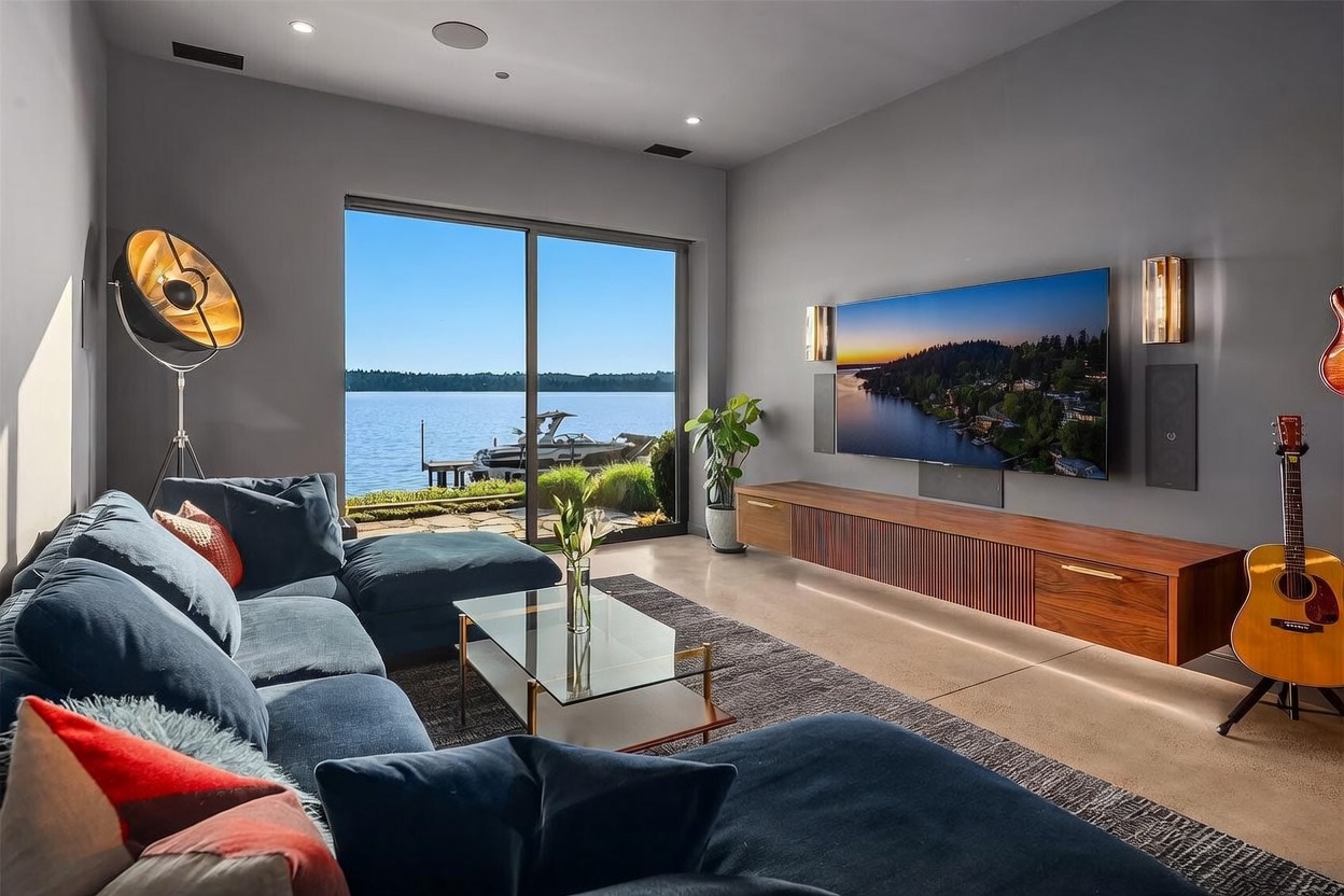 This modern living room features a large, plush blue sectional sofa and a glass-topped coffee table set against a backdrop of gray walls. A wall-mounted television sits above a long, slatted wood media console, while floor-to-ceiling sliding glass doors provide an expansive view of the lake and a private dock. The space is accented by a tall floor lamp, a guitar, and warm wall-mounted lighting, creating a sophisticated and serene atmosphere.