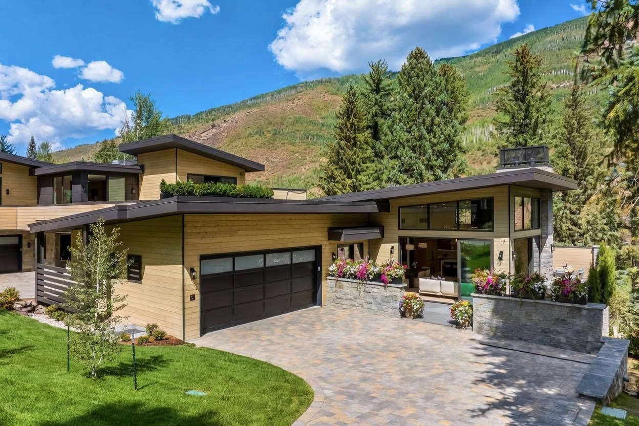 This modern mountain residence features a striking architectural design with clean lines, horizontal wood siding, and a low-pitched roof. The front exterior showcases a spacious paver driveway leading to a multi-car garage, complemented by a stone-walled garden bed filled with vibrant flowers. Large glass sliding doors open to reveal a glimpse of the interior living space, creating a seamless transition between the home and the lush, mountainous landscape.