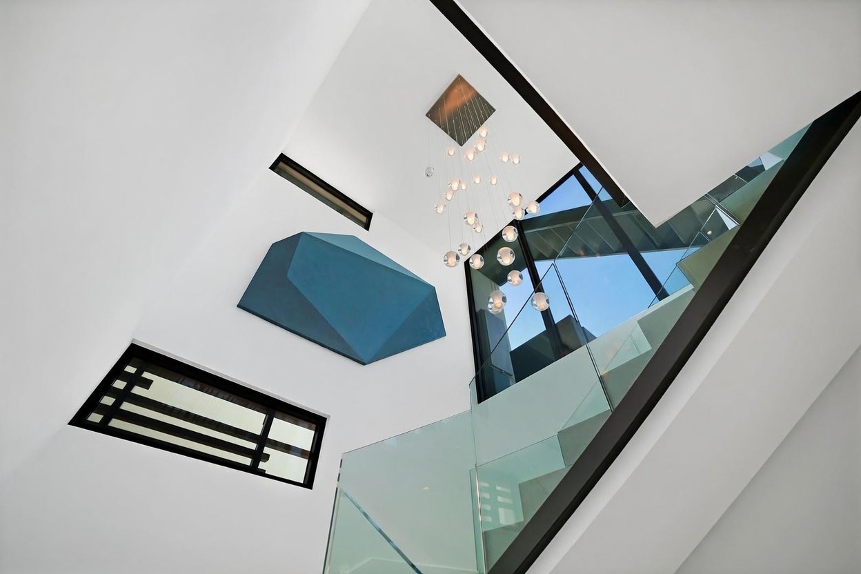 This striking interior view captures a modern staircase area characterized by a dramatic, multi-tiered glass chandelier suspended from a high ceiling. The space features a bold, geometric blue art installation mounted on a stark white wall, complemented by a rectangular window with a dark frame. A glass railing follows the curve of the staircase, emphasizing the open, airy, and contemporary architectural design of the home.