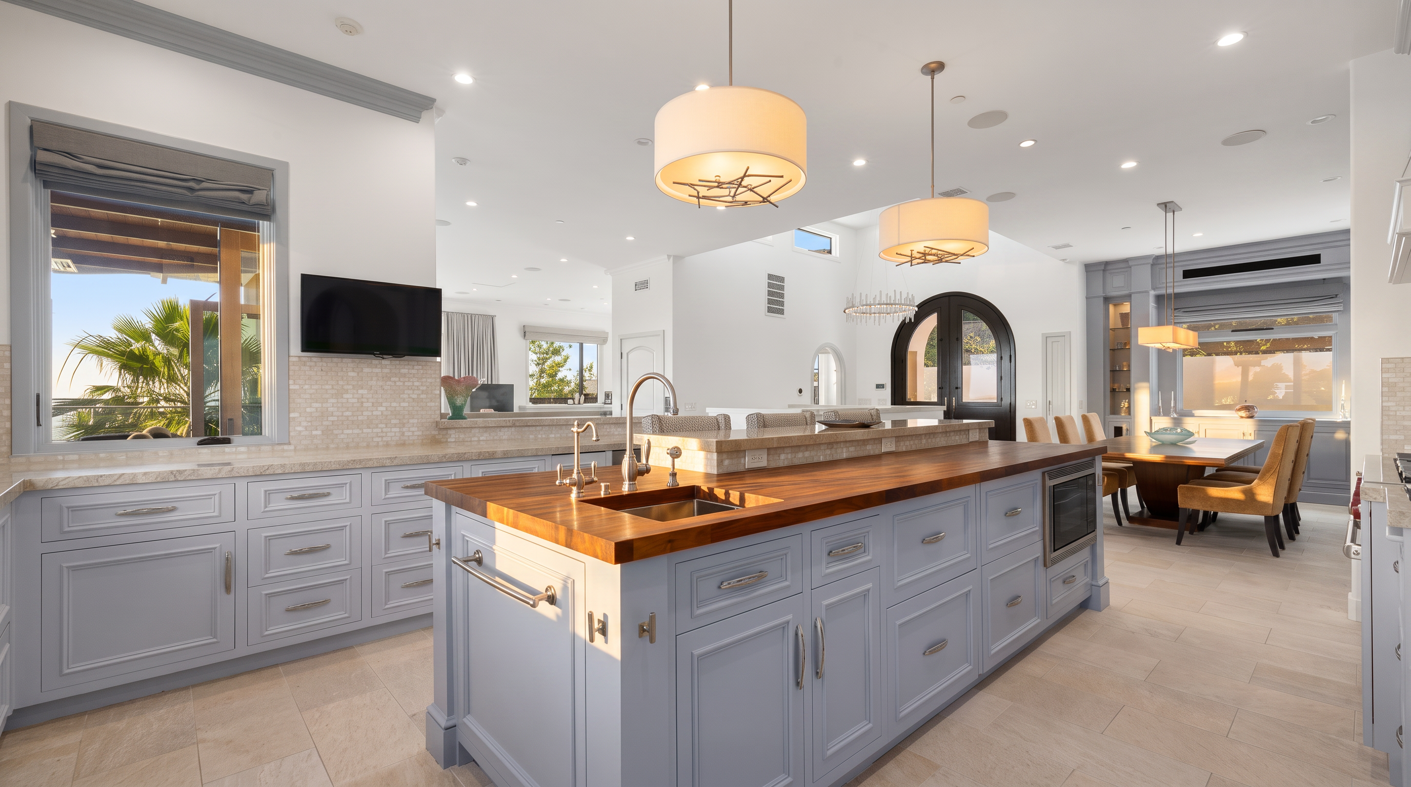 This spacious, light-filled kitchen features a large central island with a wood countertop and integrated sink, complemented by light blue cabinetry and neutral stone flooring. The open-concept design flows seamlessly into a dining area, with large windows providing natural light and views of the outdoors. Two decorative pendant lights hang above the island, creating a warm and inviting atmosphere in this well-appointed culinary space.