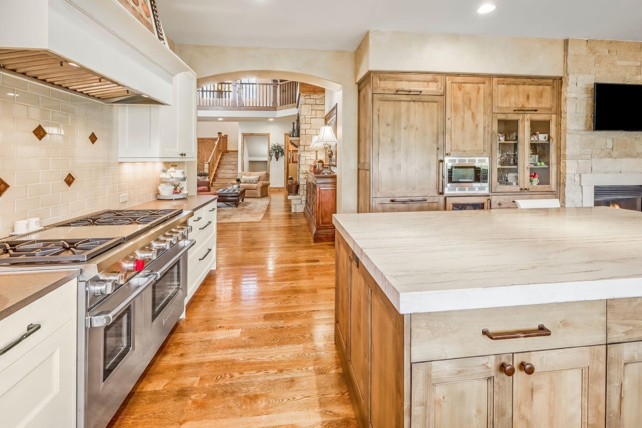 This spacious kitchen features a large central island with a light-toned stone countertop and warm wood cabinetry. To the left, a professional-grade stainless steel range is set against a white tiled backsplash, while the background reveals an open-concept layout leading toward a living area with a stone-clad chimney breast. The warm wood flooring and neutral color palette create an inviting, high-end atmosphere.