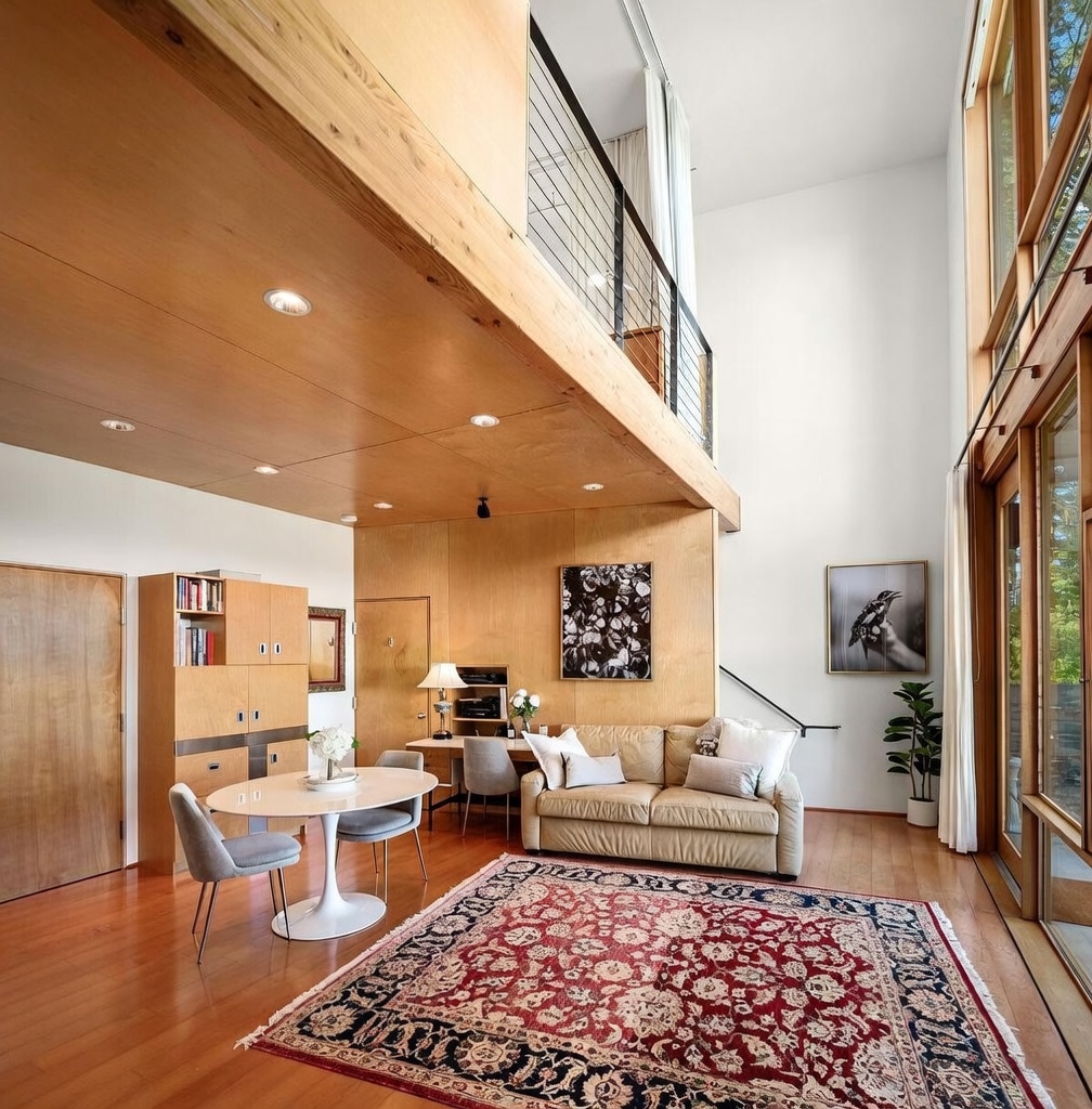 This bright, open-concept living area features a double-height ceiling with a mezzanine level overlooking the space. The room is anchored by a large, traditional patterned rug, a neutral-toned sofa, and a dining area with a modern pedestal table. Tall windows along the right wall flood the interior with natural light, highlighting the light wood paneling and warm hardwood flooring throughout.