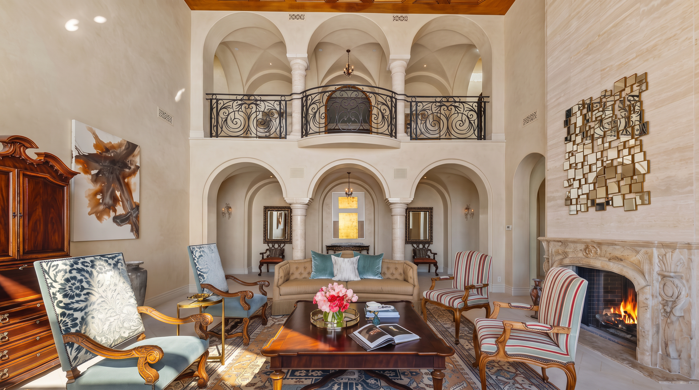 This grand, double-height living room features an ornate coffered wood ceiling and a series of arched openings that lead to an upper-level gallery with decorative iron railings. The space is anchored by a stone-clad chimney breast with a burning fireplace and a large, abstract geometric wall art piece. The room is furnished with a mix of traditional seating, including a central sofa and patterned armchairs, all arranged on a large, intricate area rug.