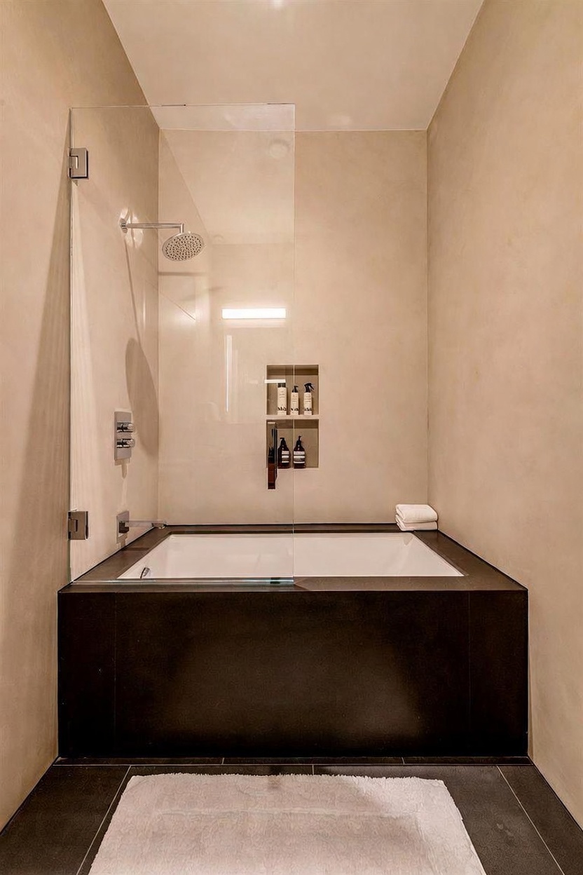 This modern bathroom features a sleek, deep soaking tub encased in a dark, matte-finished surround that contrasts with the light, neutral-toned walls. A glass partition separates the tub area from the shower, which includes a built-in recessed niche for toiletries. The minimalist design is completed by a white bath mat on the dark tiled floor, creating a clean and sophisticated spa-like atmosphere.