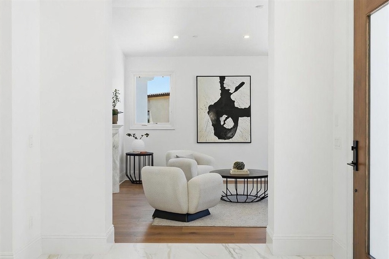 This view from a doorway captures a bright, contemporary living area featuring two curved, light-colored armchairs arranged around a circular coffee table. A large, abstract black-and-white piece of wall art serves as a focal point, while a window to the left provides natural light and a view of the exterior. The space is finished with light wood flooring and a neutral area rug, creating a sophisticated and inviting atmosphere.