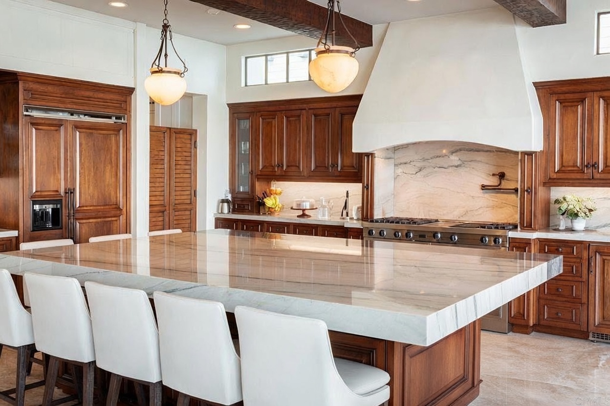 This spacious, high-end kitchen features a massive central island with a thick stone countertop and seating for several guests. The room is anchored by dark wood cabinetry and a prominent, custom-built range hood above a professional-grade stove. Warm pendant lighting hangs from the ceiling, complementing the elegant, traditional aesthetic of the space.