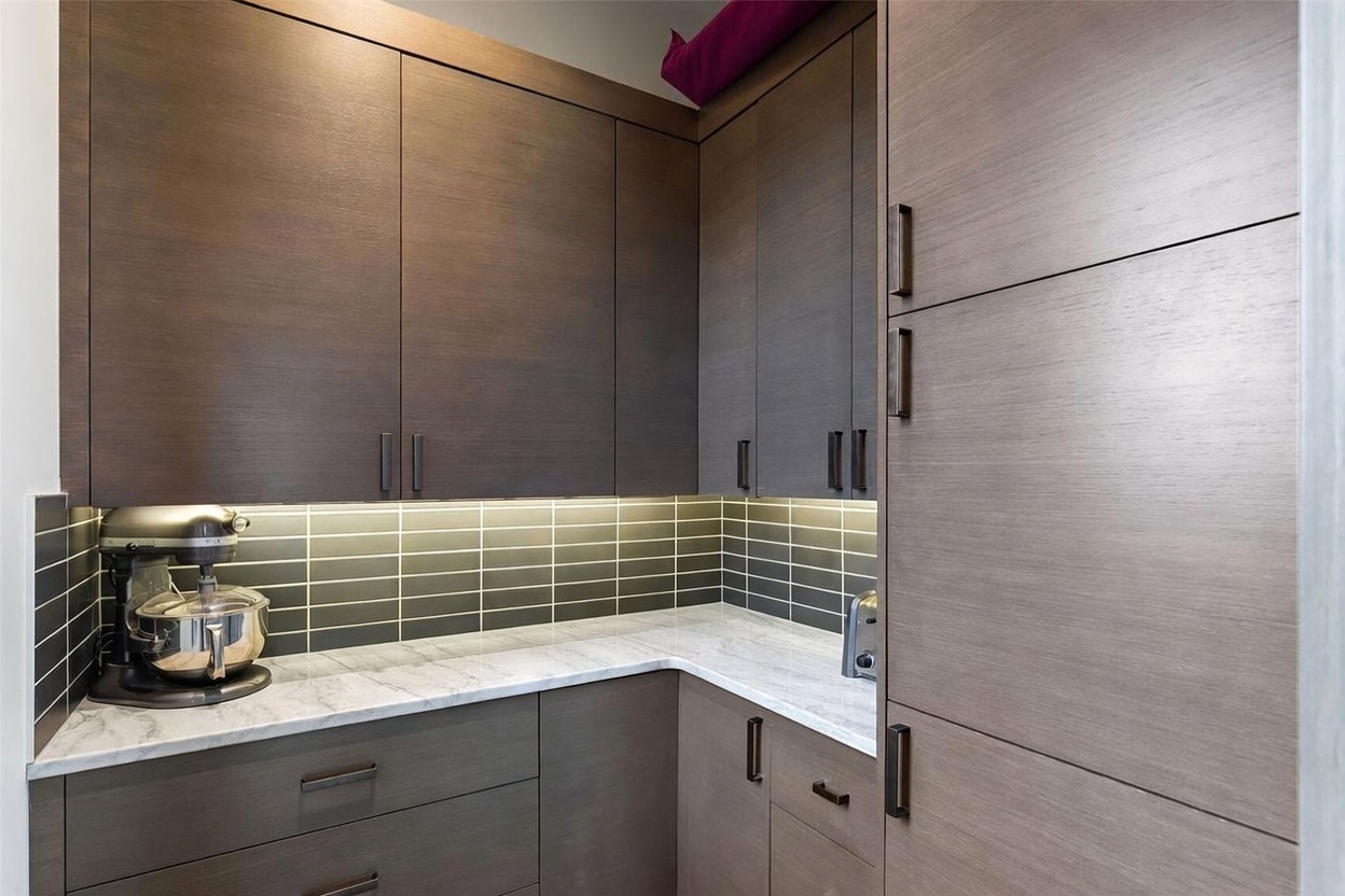 This modern kitchen area features dark, wood-grain cabinetry paired with a light-colored stone countertop and a sleek, dark-tiled backsplash. Under-cabinet lighting highlights the workspace, which includes a stand mixer and a toaster. The perspective is a close-up, eye-level shot that emphasizes the clean lines and contemporary design of the cabinetry and storage solutions.