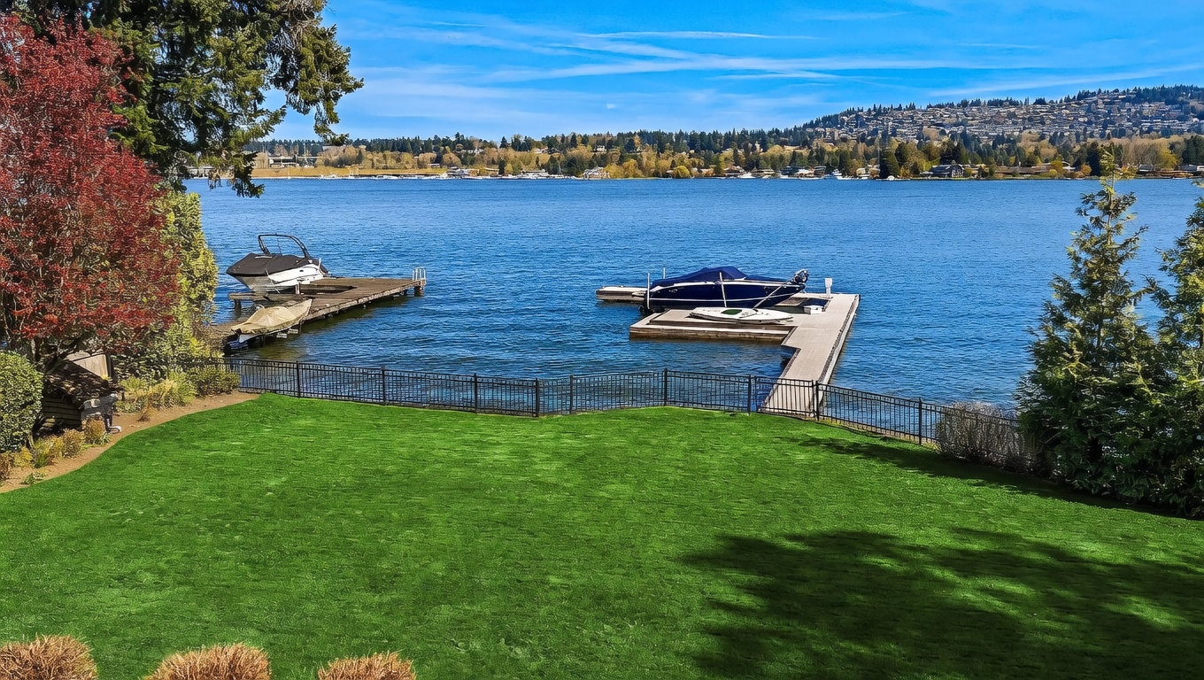 This elevated view captures a lush, manicured green lawn leading down to a private waterfront dock on a serene lake. The scene features two boats moored at the docks, framed by vibrant autumn foliage on the left and evergreen trees on the right. The perspective offers a peaceful, expansive look at the property's direct water access and scenic surroundings.