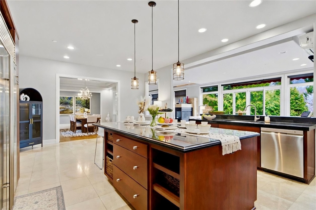 This spacious, open-concept kitchen features a large central island with a dark stone countertop and warm-toned wood cabinetry, illuminated by three industrial-style pendant lights. The space flows seamlessly into an adjacent dining area, while a wall of large windows provides ample natural light and a view of the greenery outside. The kitchen is equipped with stainless steel appliances and light-colored tile flooring, creating a bright and inviting atmosphere.