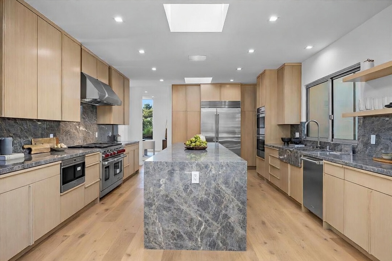 This modern, spacious kitchen features light wood cabinetry and a large central island topped with a dramatic grey stone slab. The space is illuminated by recessed lighting and two skylights, creating a bright and airy atmosphere. Stainless steel appliances, including a professional-grade range and a double-door refrigerator, are integrated into the clean, minimalist design.