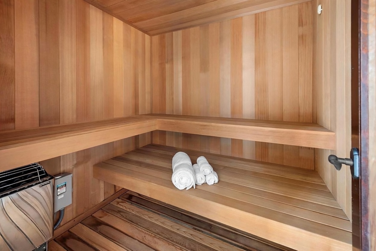 This image shows a compact, wood-lined sauna featuring tiered benches and a wall-mounted heater. The interior is constructed from light-toned wood planks, creating a warm and inviting atmosphere. A small stack of white towels rests on the lower bench, emphasizing the space's function as a private wellness amenity.