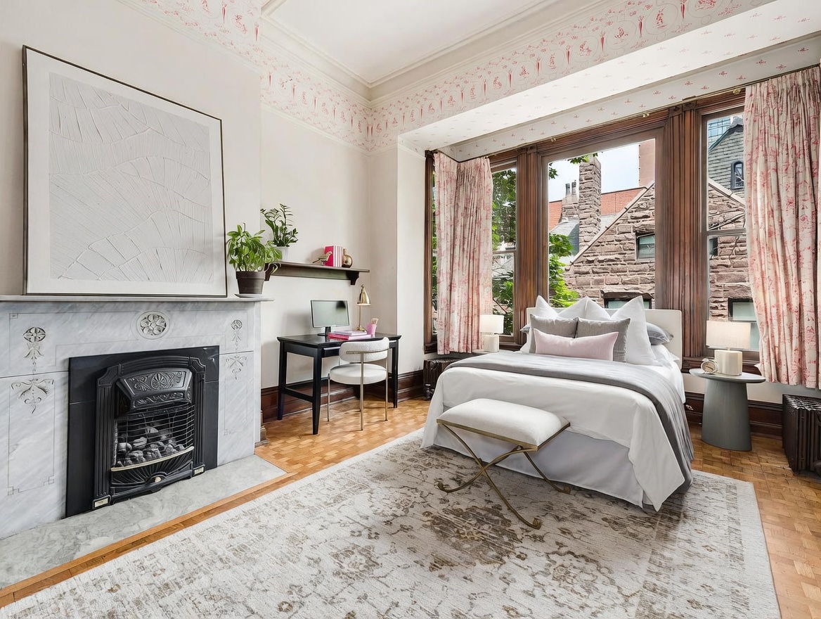 This elegant bedroom features a classic aesthetic with a prominent stone fireplace surround and a large, framed piece of abstract art above the mantel. A desk area is tucked neatly against the wall, while a bed with plush bedding sits centered in front of a large bay window framed by patterned curtains. The room is finished with warm-toned wood flooring and a decorative patterned rug, creating a sophisticated and inviting atmosphere.