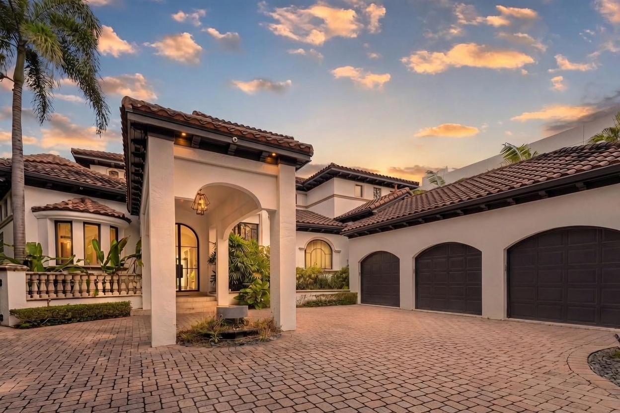 This Mediterranean-style estate features a grand, arched entryway with a decorative hanging lantern and a paved motor court leading to a three-car garage. The exterior is finished in a light stucco with a dark tile roof, complemented by lush tropical landscaping and a palm tree. The perspective is captured at dusk, highlighting the warm glow of the home's lighting against a vibrant, cloud-filled sunset sky.