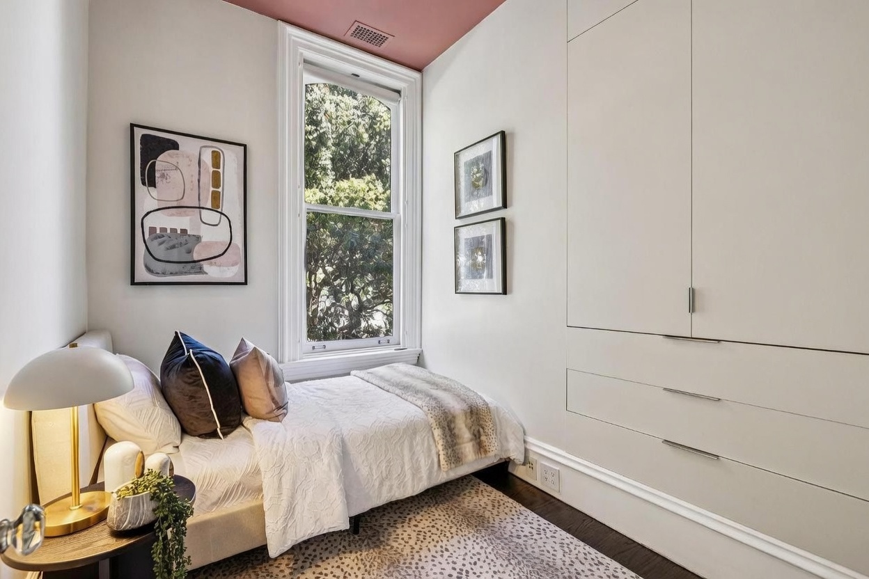 This cozy, compact bedroom features a twin bed dressed in white linens with decorative pillows, positioned next to a tall window that allows for natural light. The room is accented by a unique pink-painted ceiling, a patterned area rug, and built-in floor-to-ceiling cabinetry that provides ample storage. The space is decorated with framed artwork on the walls, creating a clean, modern, and inviting atmosphere.