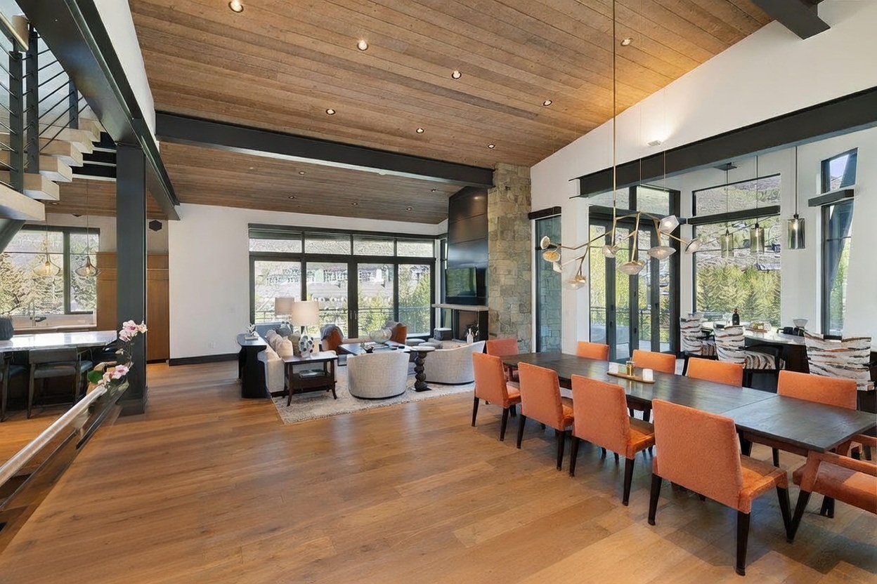 This expansive open-concept living and dining area features a high wood-paneled ceiling with recessed lighting and a striking industrial-style steel beam structure. A stone-clad chimney breast serves as a focal point between the living area and the large windows, which offer scenic views of the surrounding landscape. The space is furnished with a contemporary dining set and a comfortable seating arrangement, creating a sophisticated and airy atmosphere.